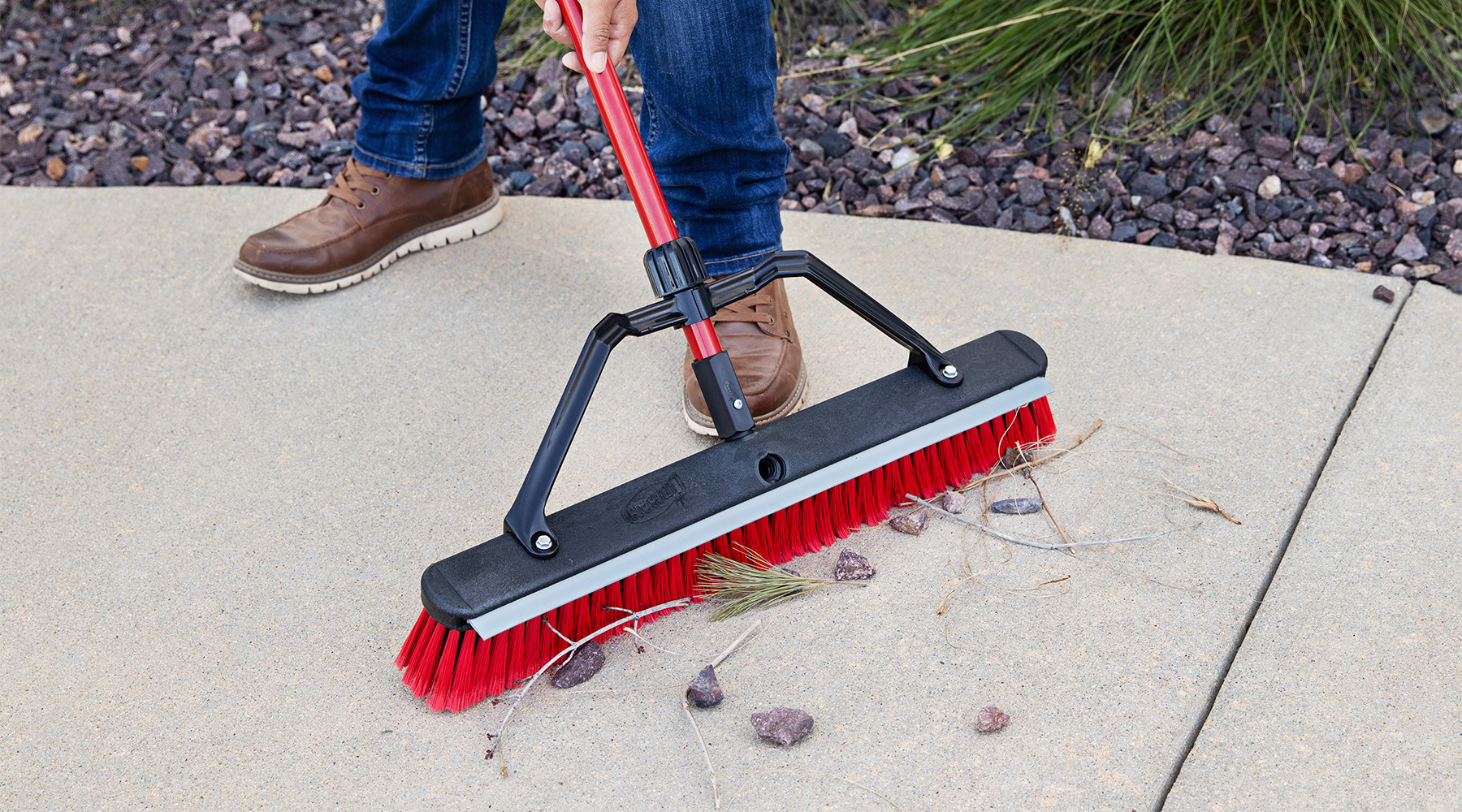 Person uses push broom to sweep rocks off of a sidewalk